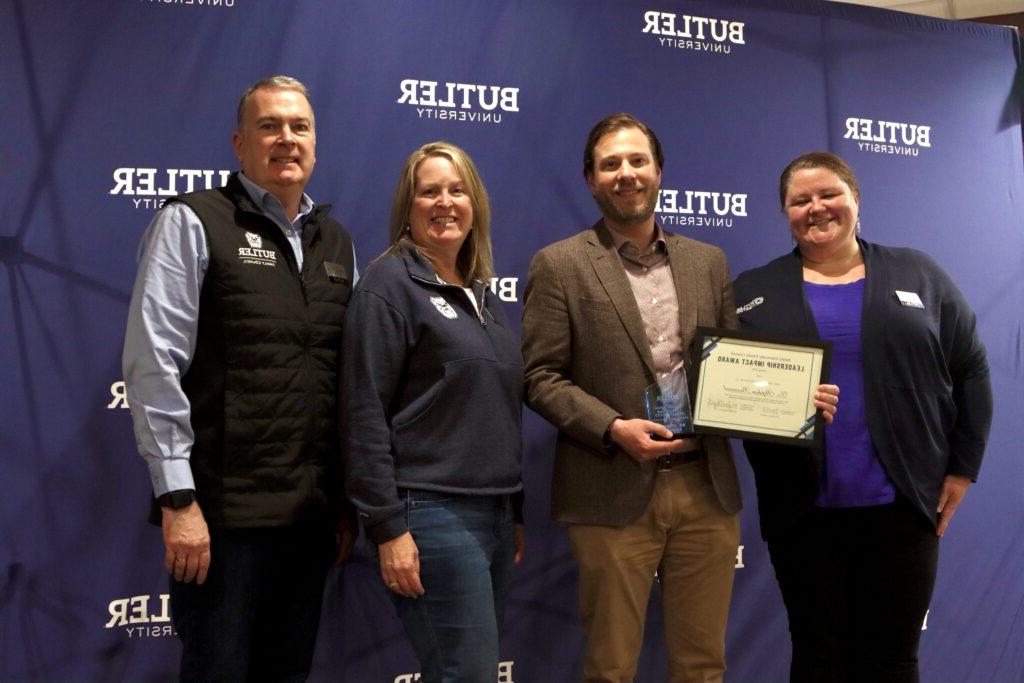 Dr. Stephen Barnard poses with his certificate and trophy alongside Director of New Student & Family Programs Meg Haggerty (left) and nominating family Jamie & David Fryrear (right). Butler University backdrop with four people posing in front. Dr. Barnard holding his certificate and trophy.