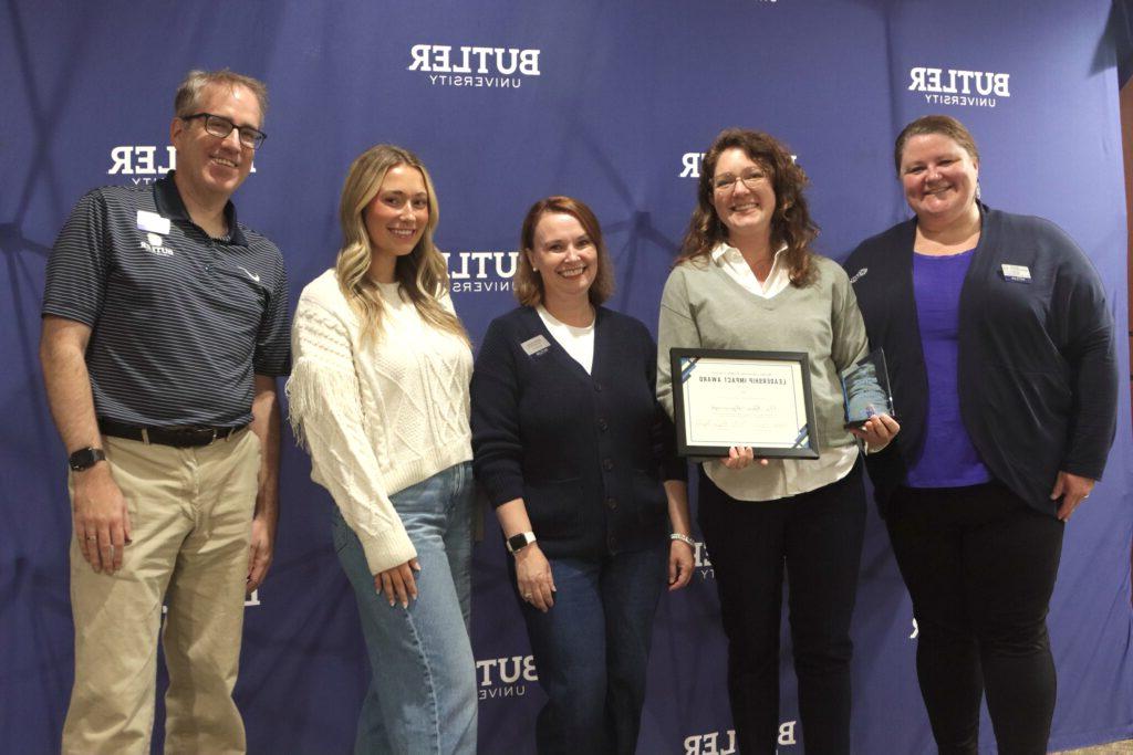 Dr. Rhea Myerscough poses with her certificate and trophy alongside Director of New Student & Family Programs Meg Haggerty (left) and nominating family Shannan & Eric Younger with student Megan McFadden (right). Butler University backdrop with five people posing in front. Dr. Myerscough holding her certificate and trophy.